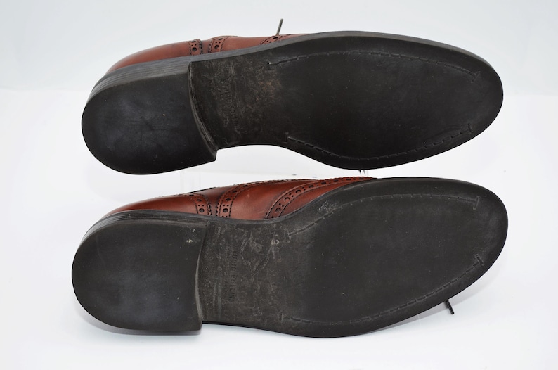 May include: A pair of brown leather shoes with a brogue design. The shoes have black soles and are shown from the bottom.