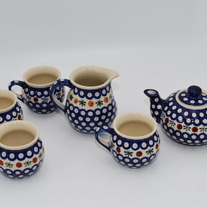 May include: A ceramic tea set consisting of a teapot, a creamer pitcher, and four mugs. Each piece features a cream-colored base with a repeating pattern of blue circles, small red dots, and green star-like shapes across the exterior surface.