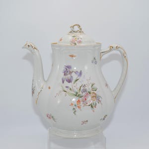 Könnte beinhalten: Eine weiße Porzellan-Teekanne mit Blumenmuster. Die Teekanne hat einen goldenen Rand und einen Deckel mit Blumenmuster. Die Teekanne ist mit verschiedenen Blumen verziert, darunter Iris, Rosen und Vergissmeinnicht.