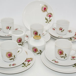 Op de afbeelding: Een wit porseleinen theeservies met een distelontwerp in roze, goud en groen. De set bevat kopjes, schotels, borden, een melkkan en een suikerpot met een gouden deksel. Het bloemmotief komt terug op alle onderdelen.