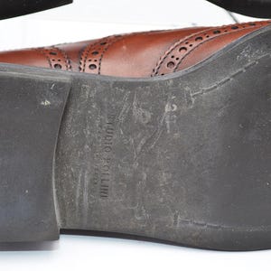 May include: Brown leather oxford shoes with a brogue design and a black rubber sole. The sole is marked with the text "Studio Pollini".