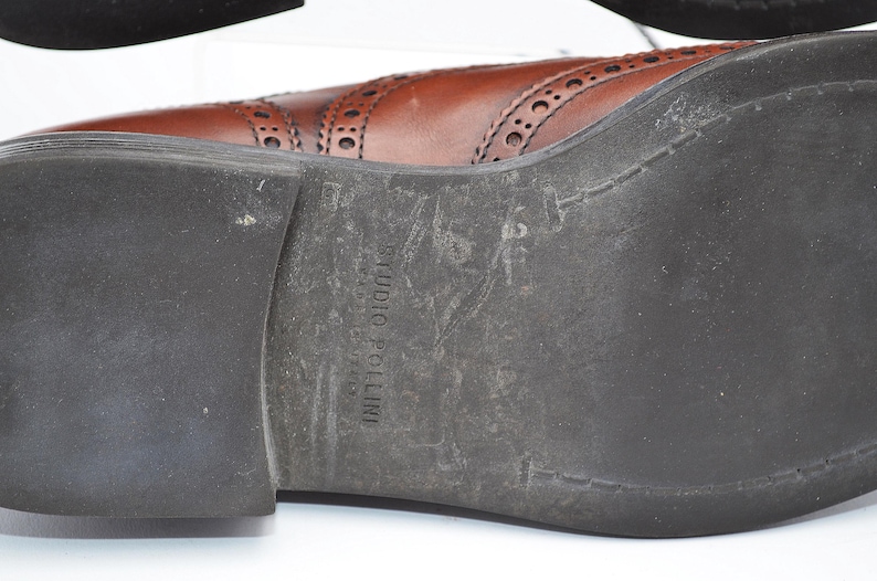 May include: Brown leather oxford shoes with a brogue design and a black rubber sole. The sole is marked with the text "Studio Pollini".