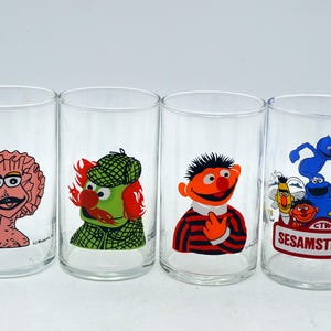 Könnte beinhalten: Ein Set aus vier klaren Trinkgläsern mit bunten Illustrationen der Sesamstraßen-Figuren. Die Gläser zeigen Charaktere wie ein rosa Wesen, ein grünes Wesen mit Hut, Ernie sowie eine Gruppe mit Bibo und dem Krümelmonster unter dem Schriftzug.