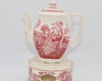 Op de afbeelding: Een crèmekleurige keramische theepot met bijpassende theelichthouder, beide versierd met een rood toile-de-jouypatroon van bomen, gebouwen en figuren. De theepot heeft een deksel, een gebogen tuit en een handvat op een ronde voet.