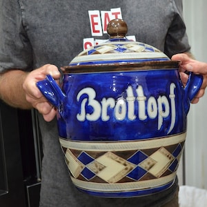 Może przedstawiać: Duży, okrągły niebieski ceramiczny pojemnik na chleb z pasującą pokrywką i dwoma bocznymi uchwytami. Z przodu znajduje się biały napis "Brottopf" stylizowaną czcionką. Podstawa i pokrywka ozdobione są geometrycznym wzorem w romby w kolorze niebieskim, brązowym i kremowym.