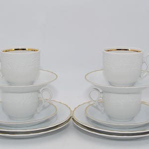 Könnte beinhalten: Zwei Sets weißer Teetassen und Untertassen mit Goldrand. Jedes Set enthält eine Teetasse, eine Untertasse und zwei zusätzliche Teller. Die Tassen und Teller haben ein zartes geprägtes Muster.