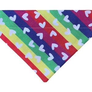 May include: Rainbow striped dog bandana with white hearts. The bandana is made of fabric and has a triangular shape.