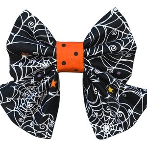 May include: A black and white spiderweb bow with orange and white polka dot accents. The bow is made of fabric and is perfect for Halloween.