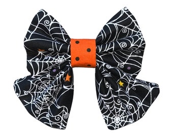 May include: A black and white spiderweb bow with orange and white polka dot accents. The bow is made of fabric and is perfect for Halloween.