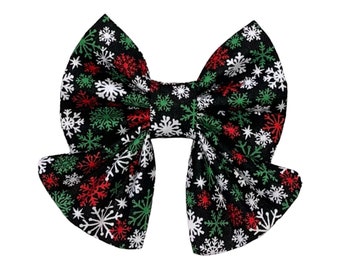 May include: A large, decorative hair bow with a black background and a pattern of white, red, and green snowflakes. The bow is tied in the center and has long, flowing tails. The bow is likely made of fabric and is suitable for the holiday season.