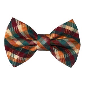 May include: A bow tie featuring a diagonal plaid pattern in shades of deep red, forest green, burnt orange, and cream. The fabric has a visible woven texture, and the bow tie is constructed with a central knot piece connecting two symmetrical wings.