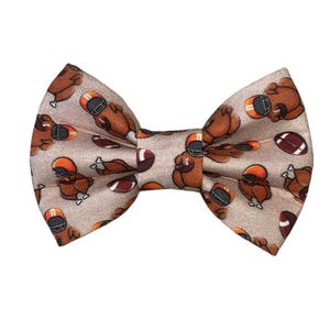 May include: A beige bow tie with a pattern of brown turkeys and footballs.
