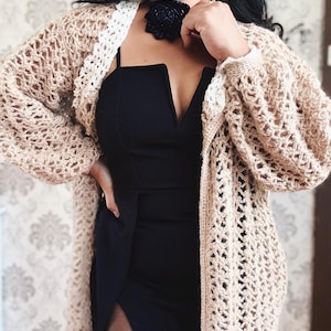 Op de afbeelding: Een vrouw draagt een zwarte jurk met een gehaakt vest. Het vest is beige en heeft een witte gehaakte rand. De vrouw draagt een zwarte bloemenketting.