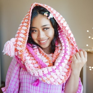 May include: A pink, white, and orange crocheted cowl with a hood and a tassel. The cowl is worn by a person with long dark hair.