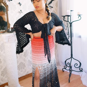 May include: A woman wearing a black crochet top with a tie front and a long, ombre crochet skirt with a slit. The skirt is a gradient of red, pink, grey, and black.