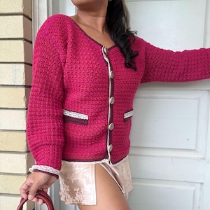 May include: A pink crocheted cardigan with white lace trim and brown buttons. The cardigan has a scoop neck and long sleeves.