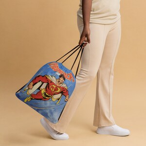 May include: A person is holding a blue drawstring bag with a comic book character design. The character is wearing a red and yellow suit and is flying through the air. The bag has the words "Superback" printed on it.