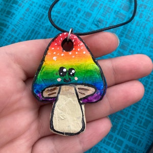 Puede incluir: Un collar con un colgante de hongo arcoíris con una cara blanca y ojos negros. El colgante está hecho de arcilla y tiene un cordón negro.