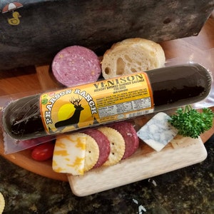 May include: A package of Pearson Ranch Venison Hickory Smoked Summer Sausage with a yellow label and a deer logo. The sausage is in a clear plastic package and is on a wooden cutting board with crackers, cheese, and parsley.