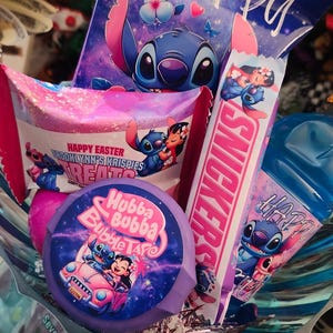 Op de afbeelding: Een cadeaupakket gevuld met lekkernijen met het personage Stitch. Het bevat een chocoladereep, Hubba Bubba Bubble Tape en zakjes snoep. De overheersende kleuren zijn blauw, roze en paars, met het woord "Happy".