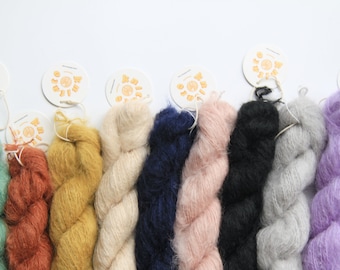 May include: Ten skeins of yarn in various colors, including teal, orange, yellow, cream, navy, pink, black, gray, lavender, and green. Each skein is wrapped in a circle and has a small tag attached with a sun logo.