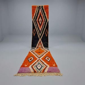 May include: A long, narrow runner rug featuring a geometric pattern of diamonds in vibrant orange, black, and purple. The design includes white and dark outlines, with a fringed edge at the bottom. The rug displays a high-pile texture throughout.