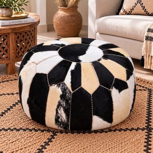 May include: A round ottoman upholstered in cowhide featuring a geometric patchwork pattern of black, white, and tan panels. The circular top and side panels are stitched together with visible light-colored thread, creating a structured, low-profile seat.