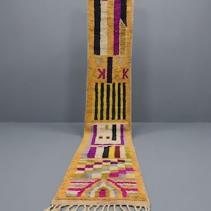 May include: A long, narrow runner rug featuring a vibrant mustard yellow base with geometric patterns in deep purple, black, white, and magenta. The design includes vertical stripes, checkered blocks, and stylized symbols, finished with cream-colored fringe.