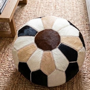 May include: A round ottoman constructed from stitched panels of hair-on hide in a geometric pattern. The surface features a central dark brown hexagon surrounded by alternating panels of black, cream, beige, and white, all joined by visible light stitching.