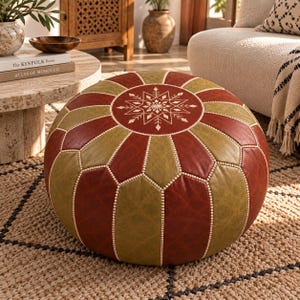 May include: A round, low-profile ottoman featuring alternating panels of deep red and olive green faux leather. The top center displays a white, intricate, geometric snowflake-like pattern, with light-colored stitching along all panel seams.