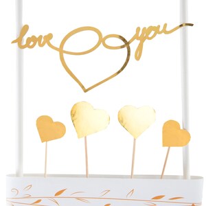May include: A white cake topper with gold detailing. The topper has the words "love you" in a cursive font, a large heart shape, and four smaller heart-shaped decorations. The base is white with gold leaf designs.