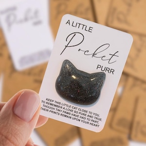 May include: A black cat-shaped charm with a glittery finish attached to a white rectangular card. The card features the text A LITTLE Pocket PURR and a poem that reads KEEP THIS LITTLE CAT CLOSE TO YOU, TO REMEMBER A LOVE SO PURE AND TRUE.