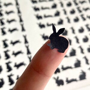 Puede incluir: Un recorte de papel negro de un conejo en la punta de un dedo. El fondo es una hoja de papel con muchos recortes de papel negro m&aacute;s peque&ntilde;os de conejos.