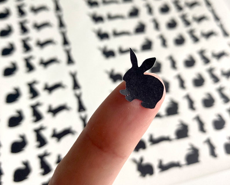 Puede incluir: Un recorte de papel negro de un conejo en la punta de un dedo. El fondo es una hoja de papel con muchos recortes de papel negro m&aacute;s peque&ntilde;os de conejos.