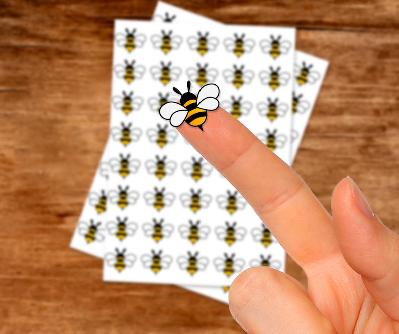May include: A sheet of white paper with multiple black and yellow bee stickers. A finger is holding one of the bee stickers.