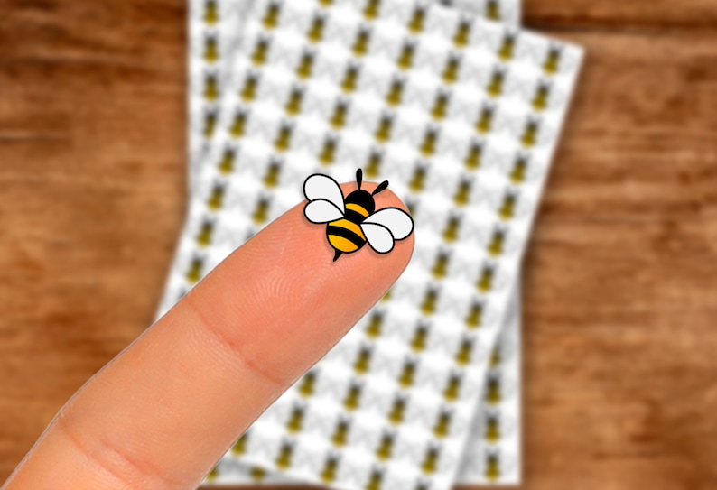 May include: A small yellow and black bee sticker on a finger. The sticker has white wings and a black head.