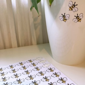 May include: A sheet of white paper with multiple black and yellow bee stickers. The bees have black eyes and wings. The stickers are arranged in rows and columns. There are four bee stickers on a white ceramic pot with green leaves.