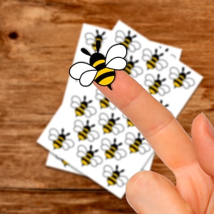 Può includere: Un foglio di adesivi con più immagini di api gialle e nere. Un dito tiene uno degli adesivi di api.