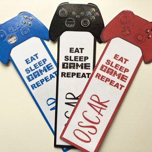 May include: Three colourful bookmarks shaped like video game controllers. The bookmarks have the text "Eat Sleep Game Repeat" and the names "Oscar" and "And" on them.