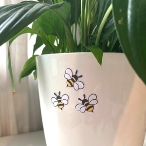 May include: A white ceramic planter with three bee stickers on the side. The bees are black and yellow with white wings.