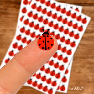 May include: A sheet of glossy stickers featuring a red ladybug design. The stickers are arranged in rows on a white background. The word "GLOSSY" is printed in black at the bottom of the sheet.