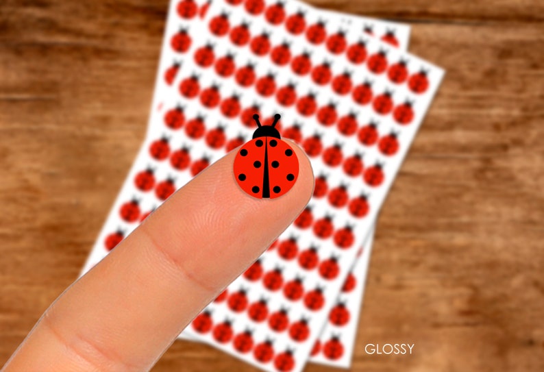 May include: A sheet of glossy stickers featuring a red ladybug design. The stickers are arranged in rows on a white background. The word "GLOSSY" is printed in black at the bottom of the sheet.