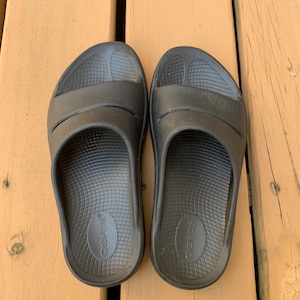 May include: A pair of black slide sandals with a contoured footbed and two straps across the top. The sandals are made of a rubber-like material and are designed for comfort. The brand name "OOFOS" is visible on the footbed.