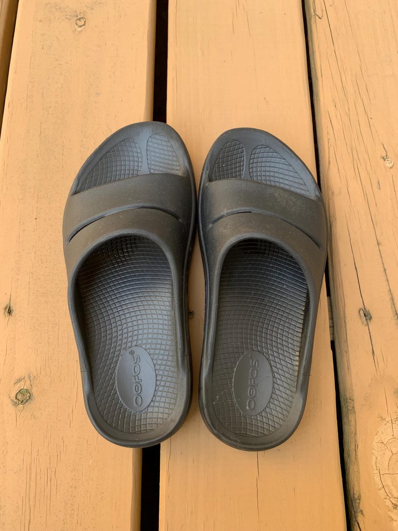 May include: A pair of black slide sandals with a contoured footbed and two straps across the top. The sandals are made of a rubber-like material and are designed for comfort. The brand name "OOFOS" is visible on the footbed.