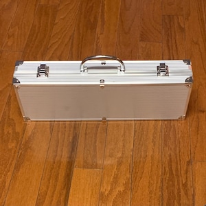 May include: A rectangular silver aluminum carrying case with a ribbed texture, featuring a central metal handle, two silver-toned locking latches, and reinforced metal corner protectors on all edges for added durability and secure storage.