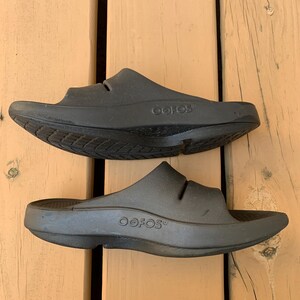 May include: A pair of black OOFOS slides, viewed from above. The slides are made of a solid, matte black material. The brand name "OOFOS" is visible on the side of each slide. The slides are designed with a contoured footbed and a single strap.
