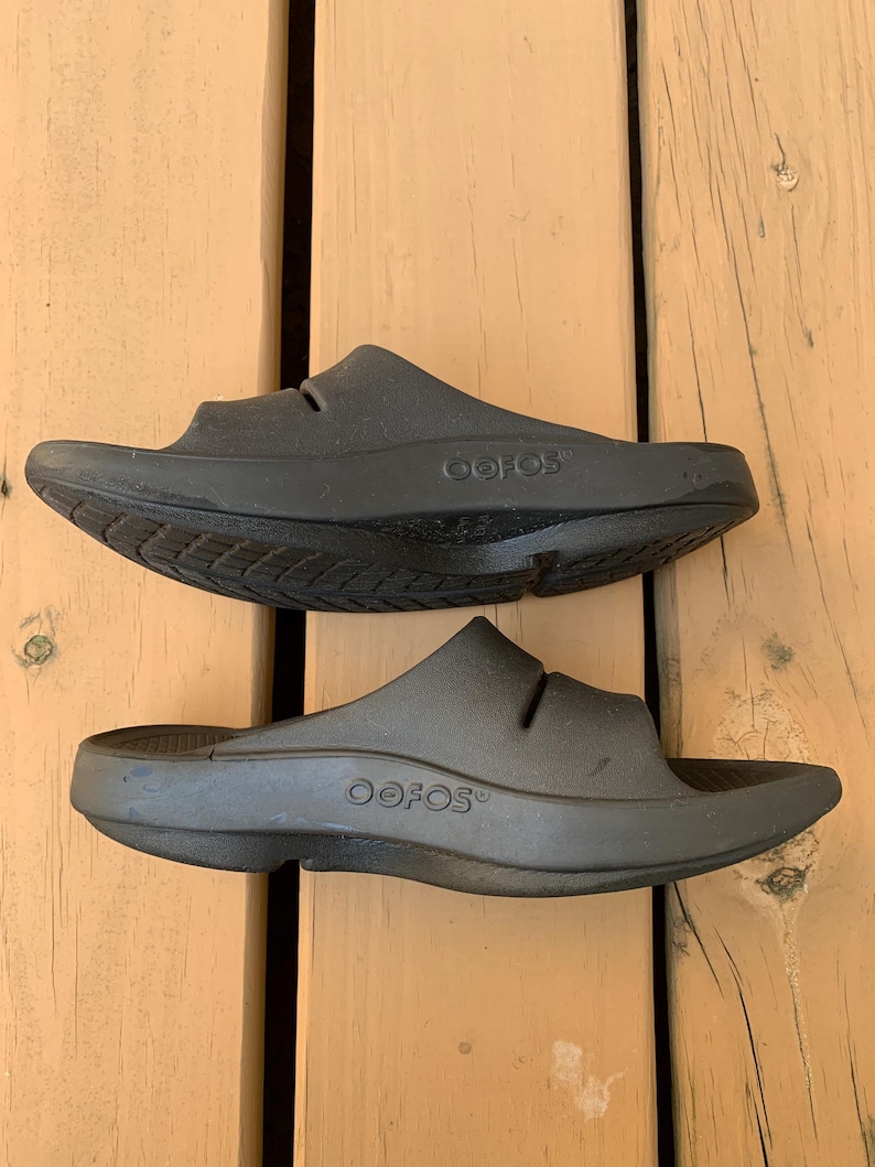 May include: A pair of black OOFOS slides, viewed from above. The slides are made of a solid, matte black material. The brand name "OOFOS" is visible on the side of each slide. The slides are designed with a contoured footbed and a single strap.