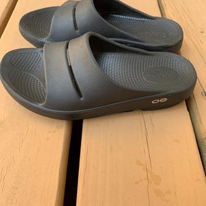 May include: A pair of black slide sandals with a contoured footbed and a wide strap across the top. The sandals are made of a rubber-like material and are designed for comfort and support. The sandals are shown on a wooden surface.