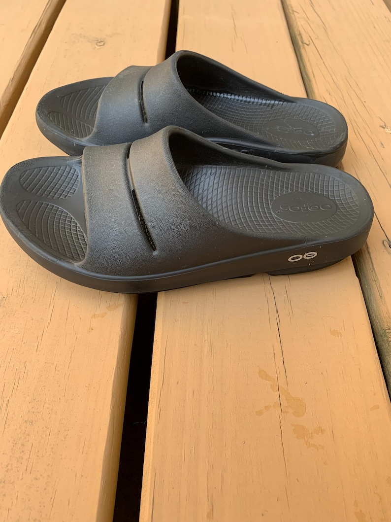 May include: A pair of black slide sandals with a contoured footbed and a wide strap across the top. The sandals are made of a rubber-like material and are designed for comfort and support. The sandals are shown on a wooden surface.