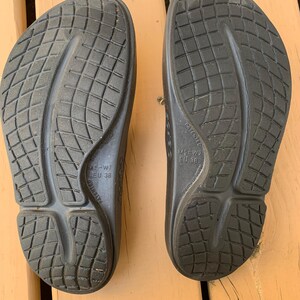 May include: A pair of black sandals, viewed from the soles. The sandals have a textured, grid-like pattern and a contoured design. The size is M5-W7, EU 38. The sandals are on a wooden surface.